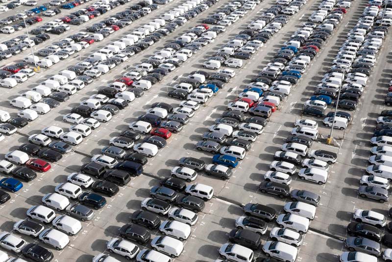 Vogelperspektive auf einen belebten Parkplatz, der mit Reihen geparkter Autos in verschiedenen Farben, überwiegend Schwarz und Weiß, gefüllt ist.