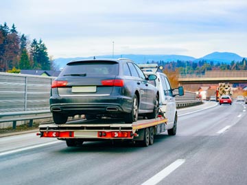 Ein Abschleppwagen von Unfallwagen Duderstadt transportiert ein schwarzes Auto auf einem flachen Anhänger entlang einer Autobahn, mit Bäumen und Bergen im Hintergrund.